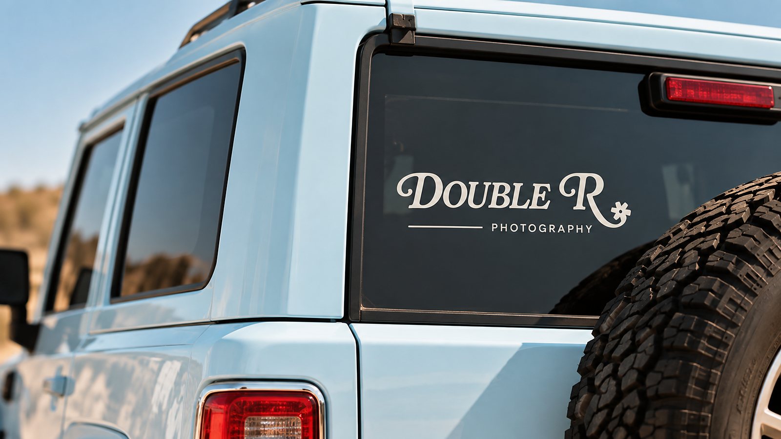 Direction Two car window decal — the cream script Double R Photography wordmark applied to the back window of a sky-blue Jeep, warm daylight.