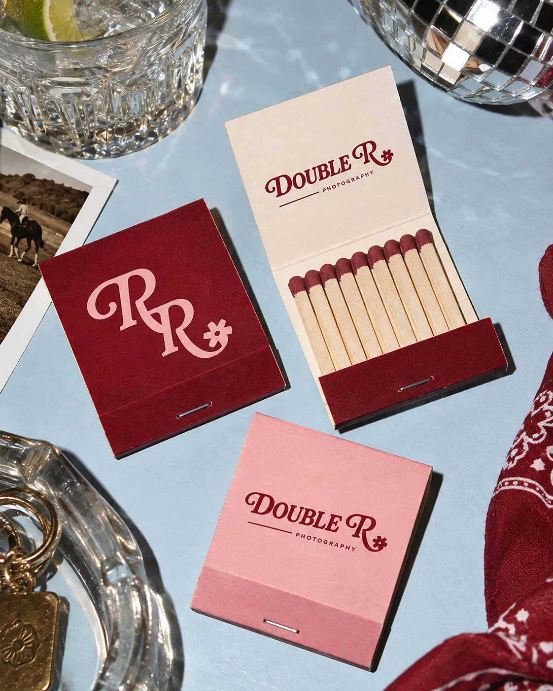 Direction Two matchbook mockups on a pale blue surface — a burgundy matchbook with pink RR monogram, a cream matchbook opened to show matches in burgundy, and a pink matchbook with the script Double R wordmark.